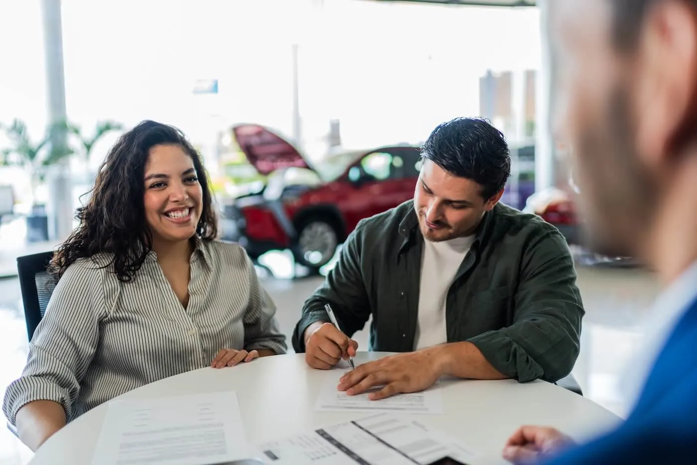 Happy couple seated at a desk in a car dealership, reviewing and signing paperwork with a sales representative. Documents are spread across the table, and vehicles are visible in the showroom background.