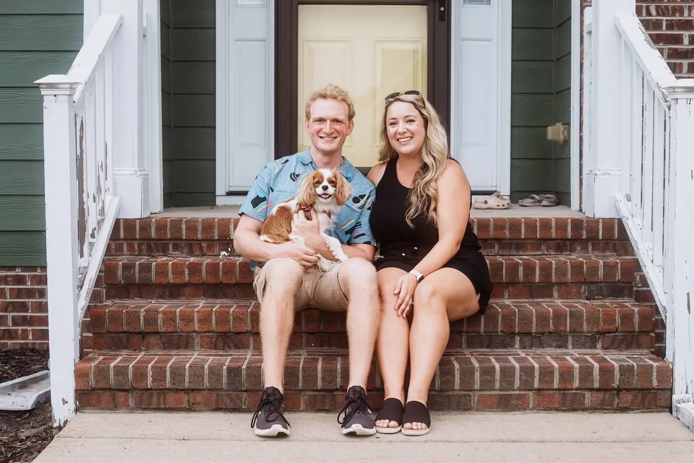 Happy couple sitting on the brick front steps of a house with a small dog, posing in front of the home's entrance and porch railing.