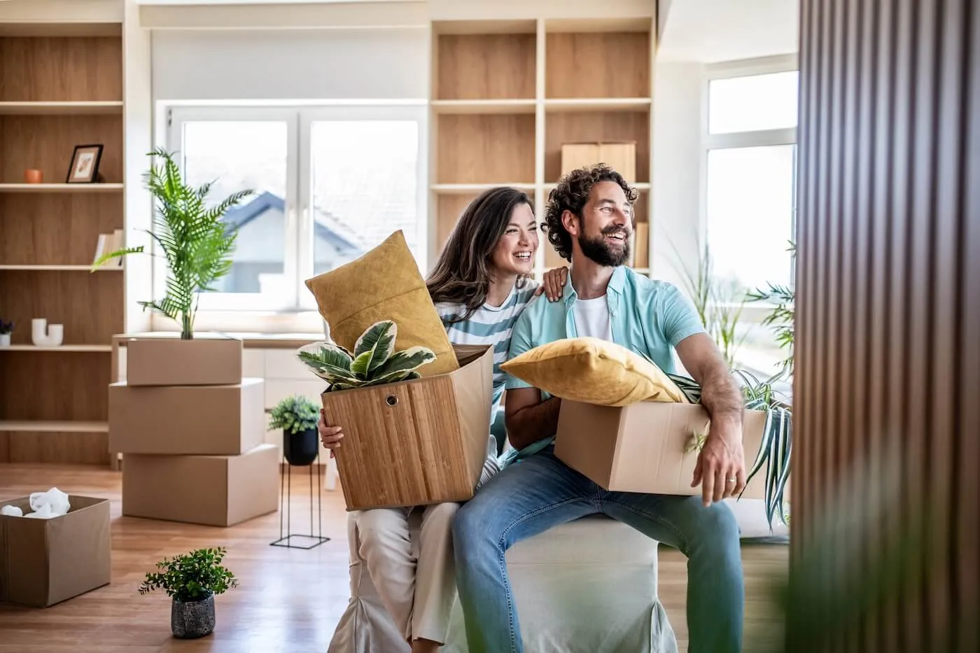 Happy couple sitting among moving boxes and household items in a bright living room, holding plants and decorative pillows while settling into a new home.