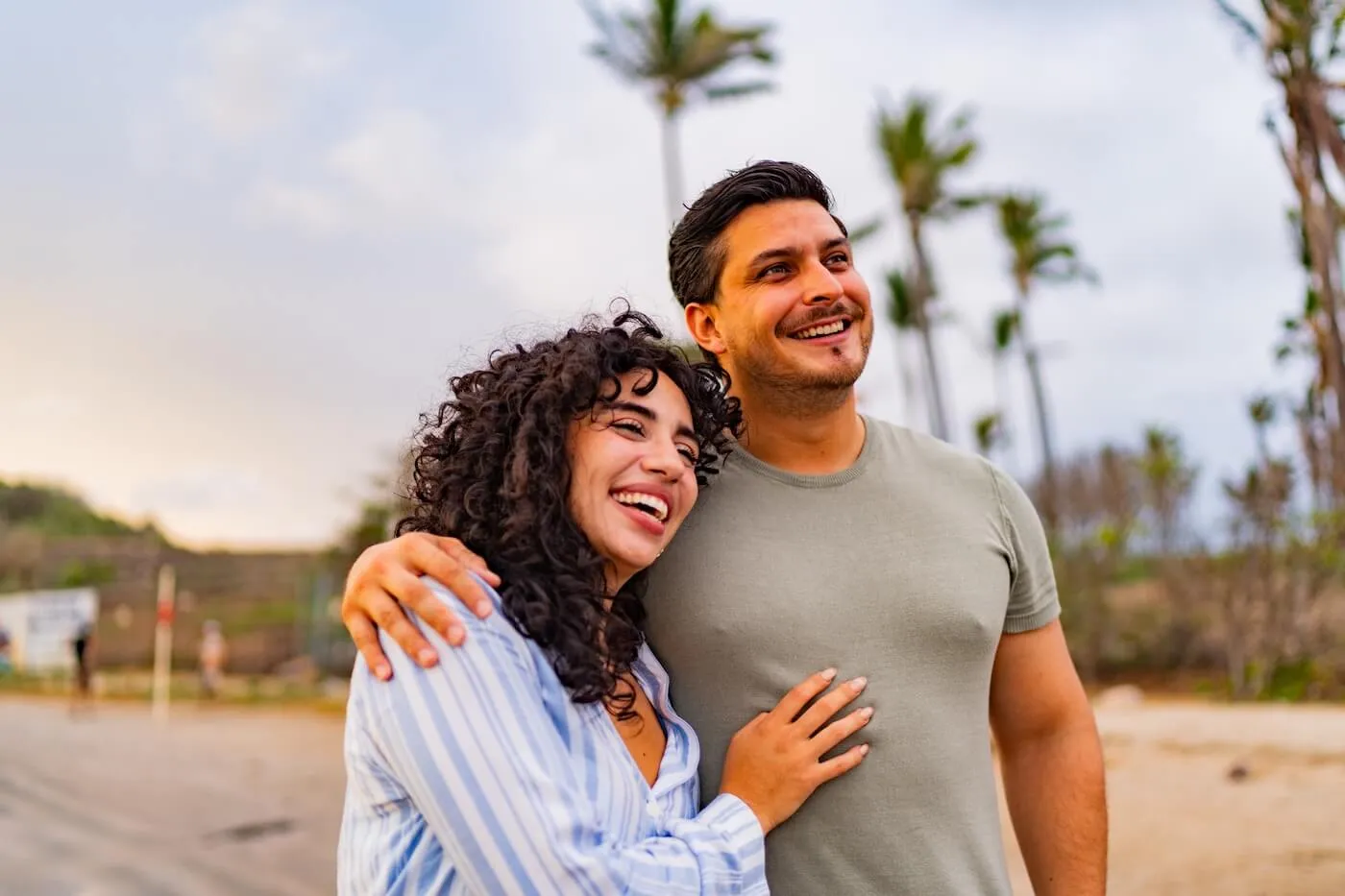 Happy couple walking together on a sandy beach with palm trees in the background, outdoors near the shoreline during daylight.