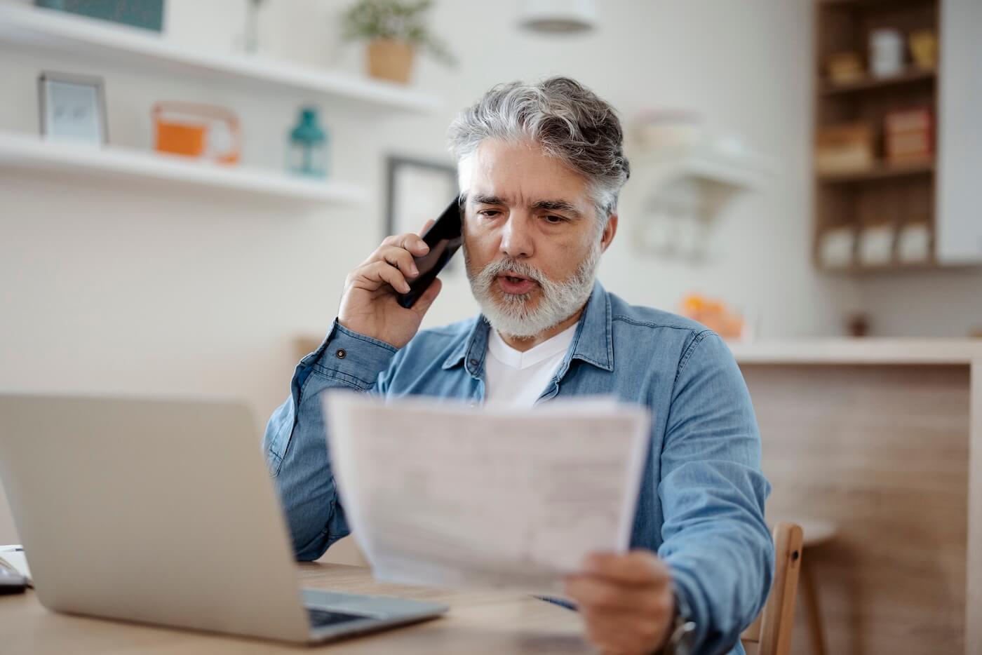 Focused senior man making a phone call while holding printed documents, with an open laptop on the desk