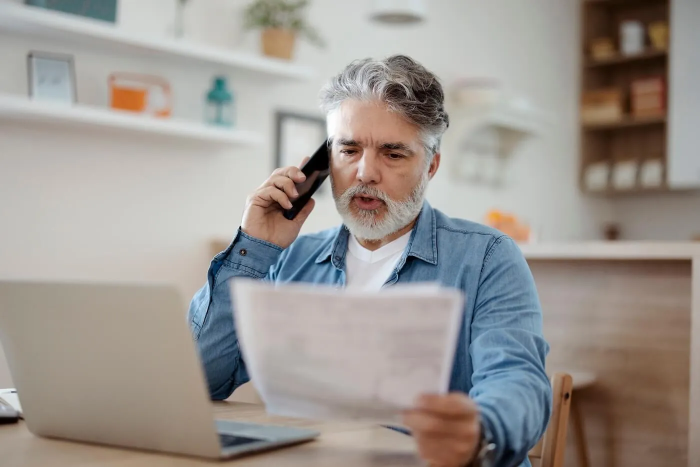 Focused senior man making a phone call while holding printed documents, with an open laptop on the desk