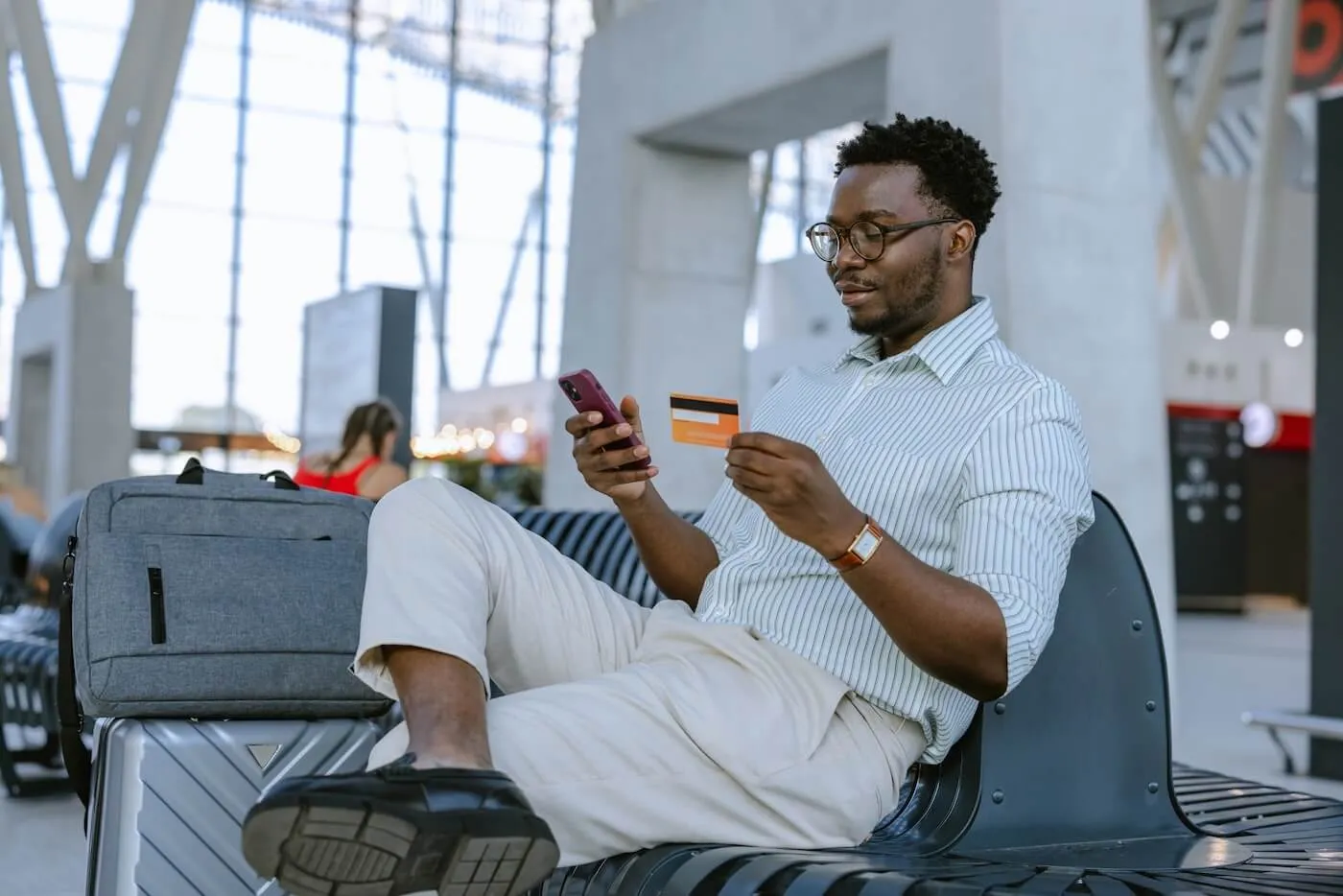 Smiling man sitting in an airport terminal, holding a smartphone in one hand and a credit card in the other, with a suitcase and backpack nearby.