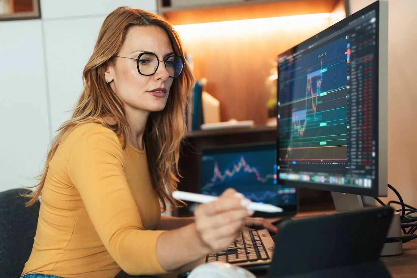 Woman working at a desk with multiple monitors displaying financial charts, using a stylus and keyboard
