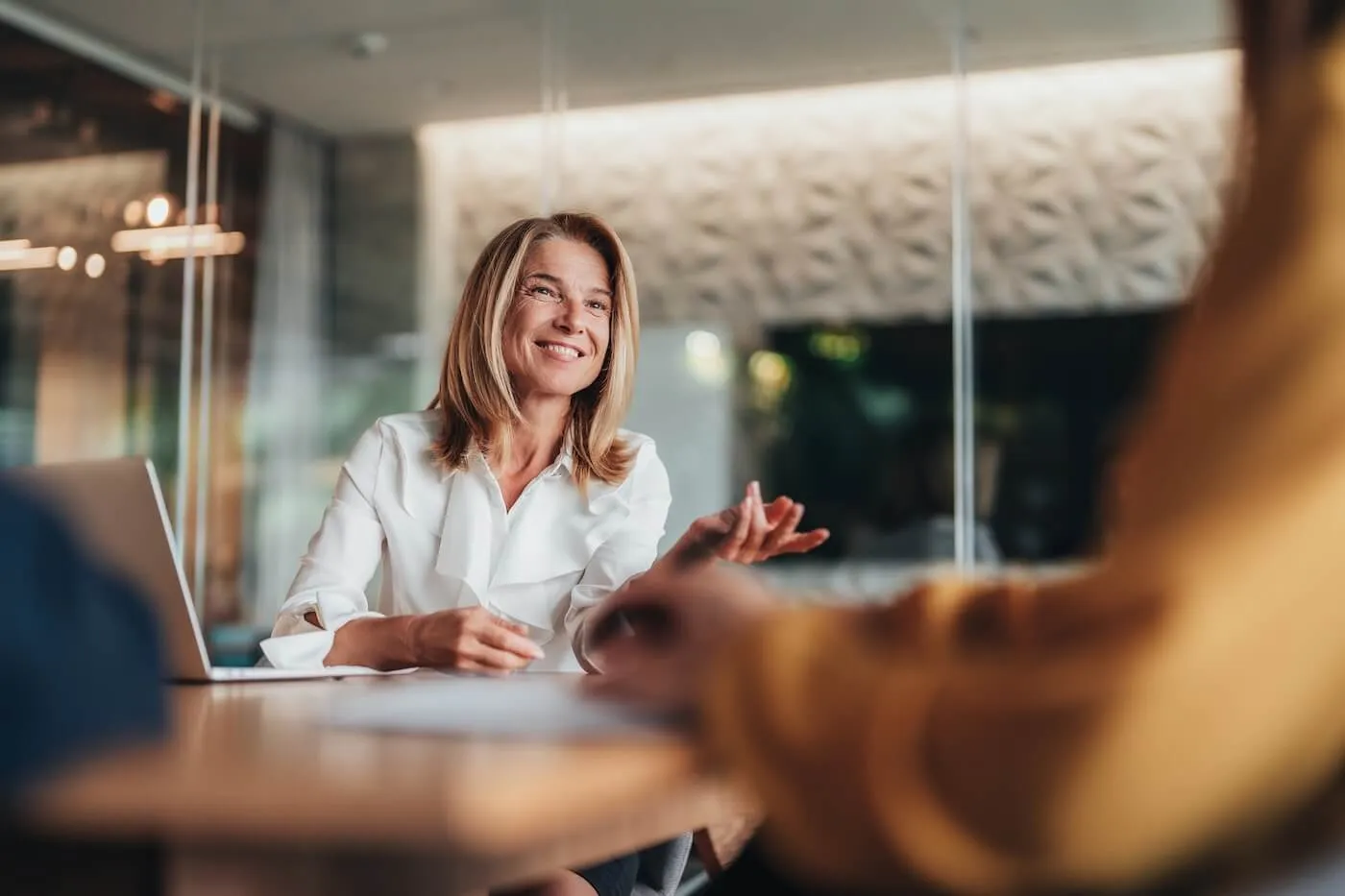 Smiling mature businesswoman sits at a table in a modern office, gesturing while speaking to someone across the desk, with a laptop open nearby.