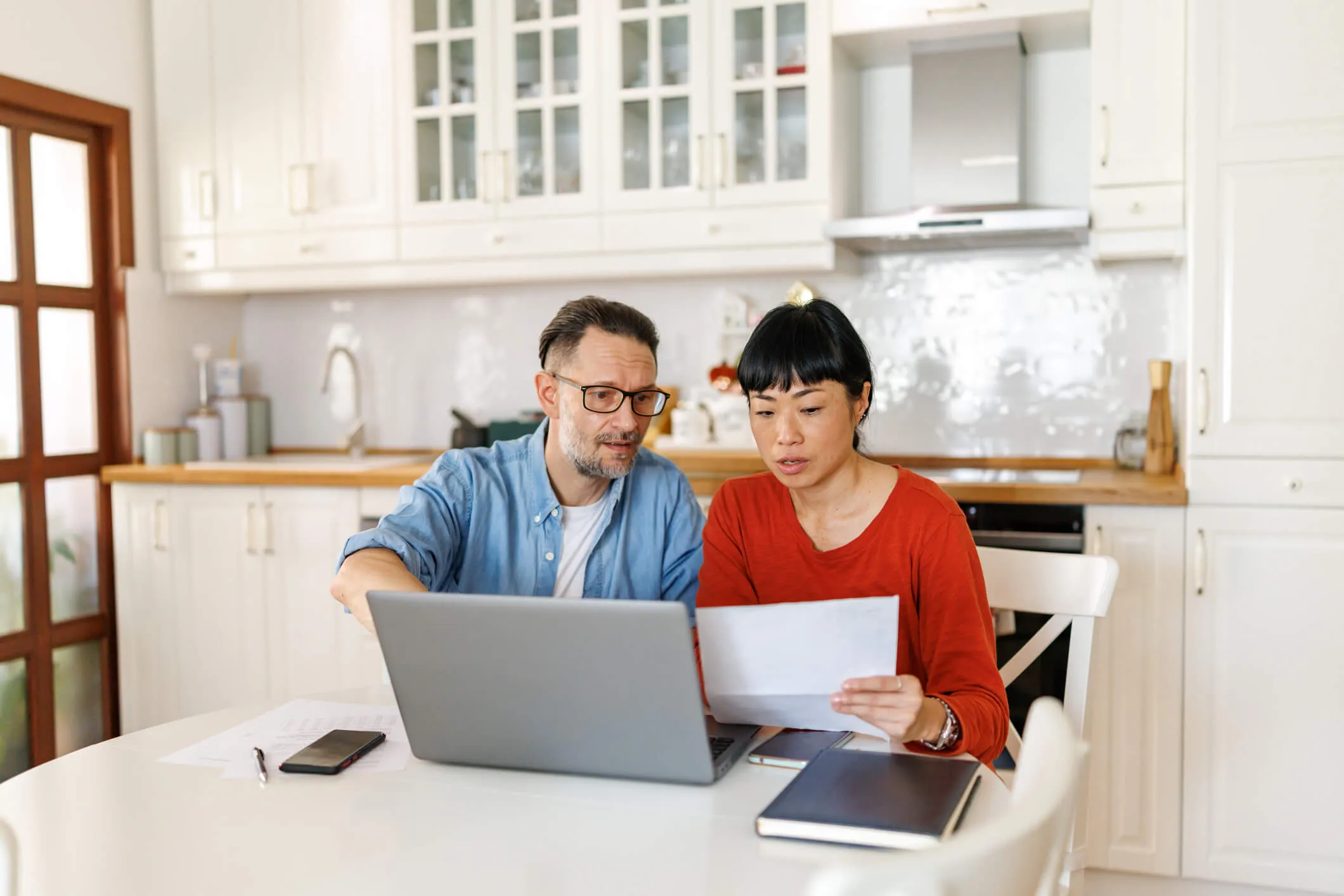 Focused mature couple sitting at dinning table and using laptop while reviewing paper financial statements at home