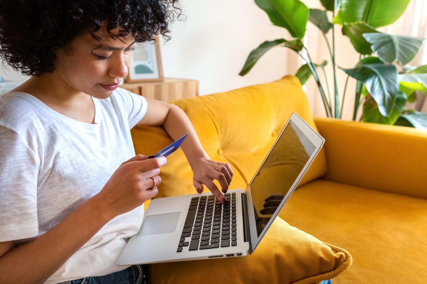 Focused woman sitting on a yellow sofa with a laptop and holding a credit card