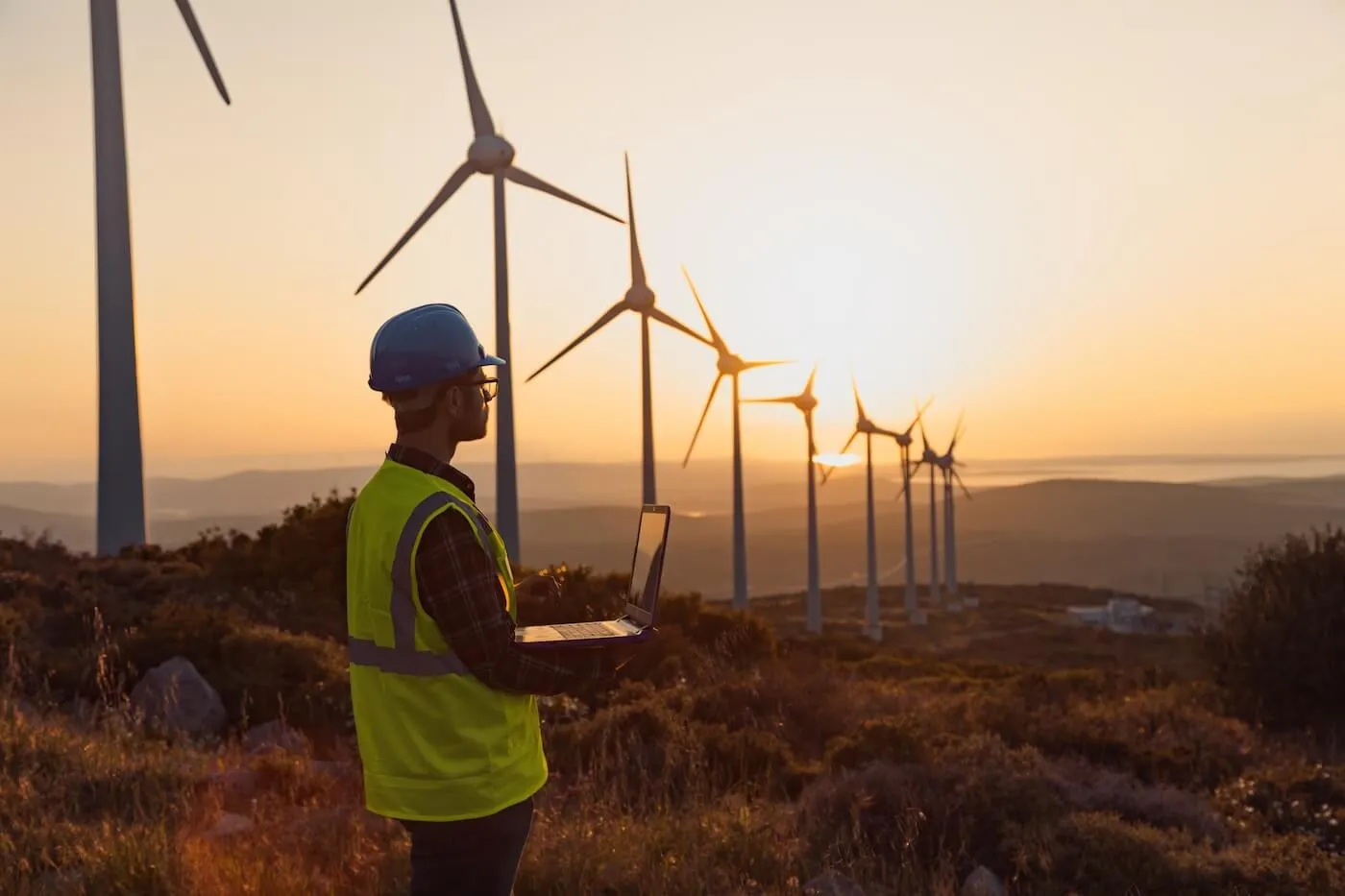 A person wearing safety gear stands in a field of wind turbines while using a laptop at sunset