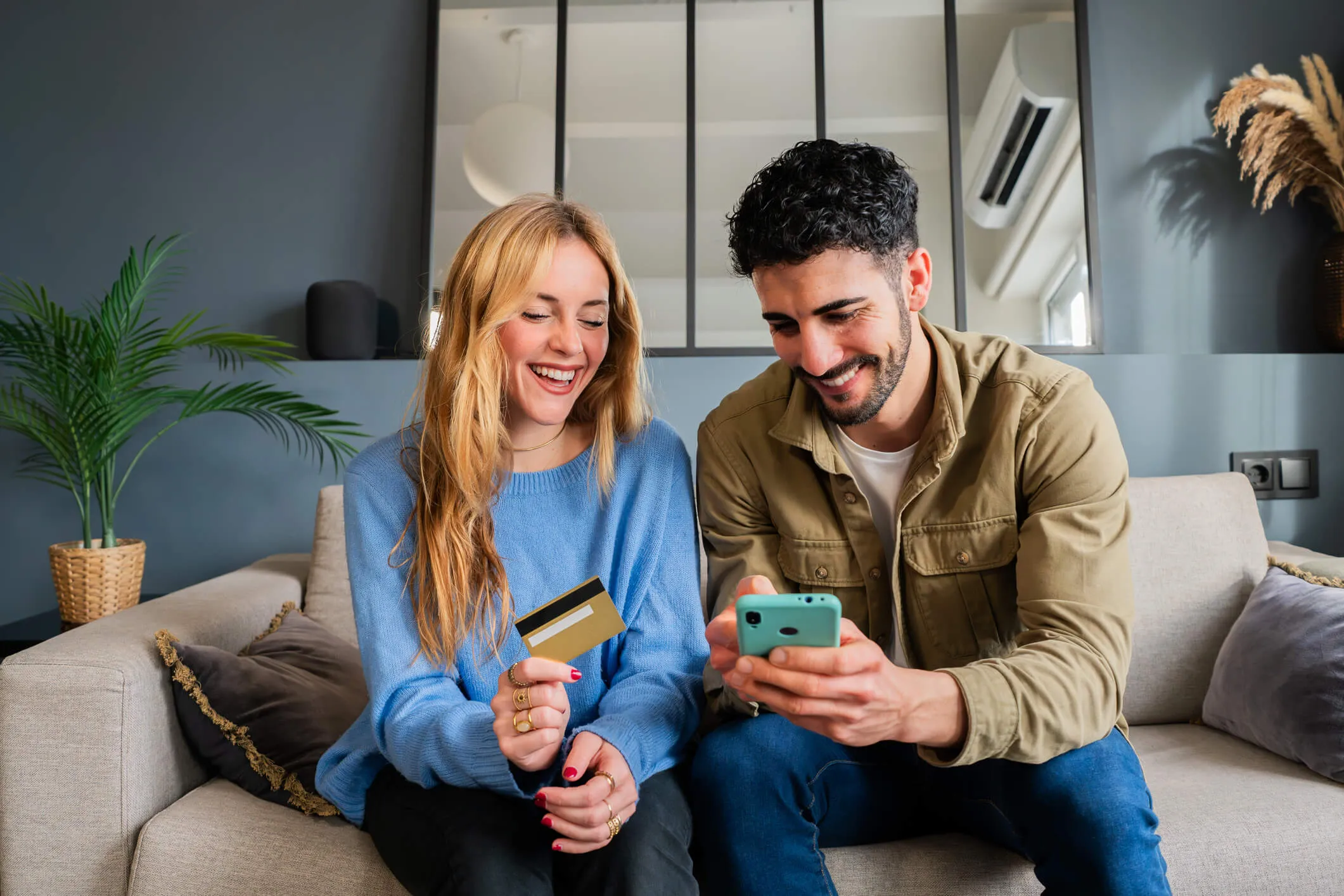 Happy couple sitting on a couch holding a smartphone and a payment card, with a coffee table and home furnishings visible nearby.