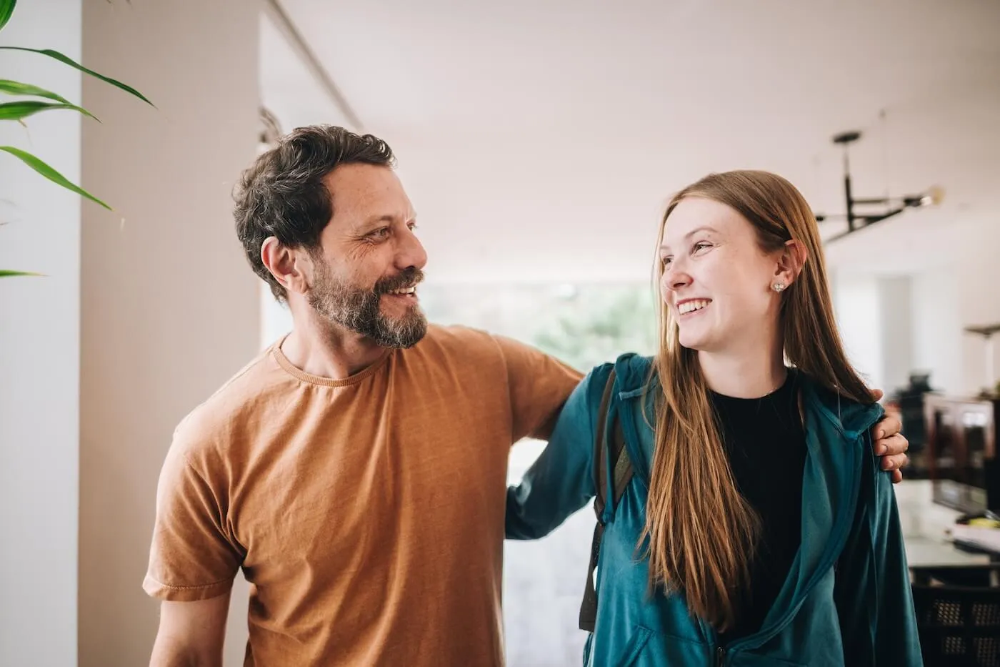 A father and a teenage daughter standing together in a bright home interior, with an arm around the teenager's shoulders and both looking toward each other near a large window.