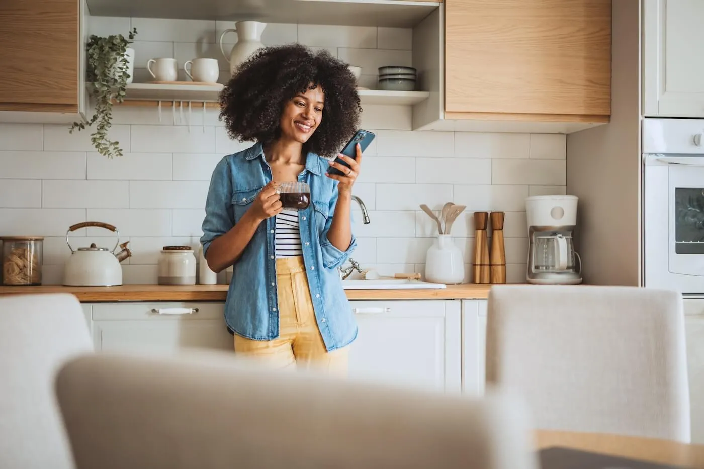 Smiling woman using her smartphone in the kitchen while holding a coffee mug