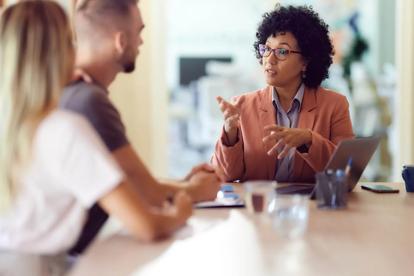 Female financial advisor seated at a conference table speaking with two clients during a consultation. A laptop, smartphone, notebooks, and glasses of water are on the table in a modern office setting.