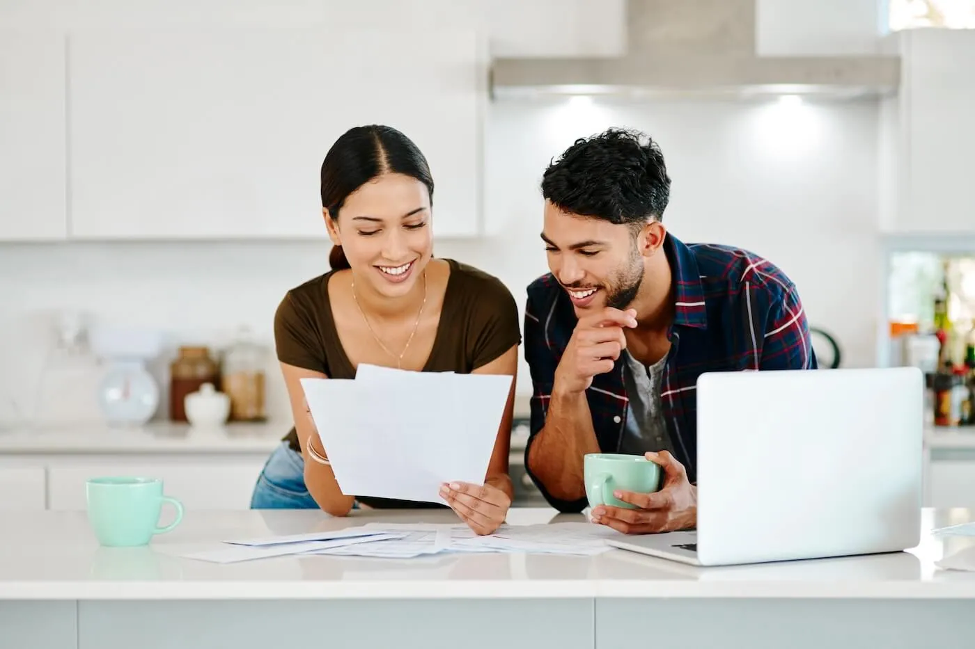 Happy couple reviewing printed documents at the kitchen counter, with an open laptop and a mug beside them