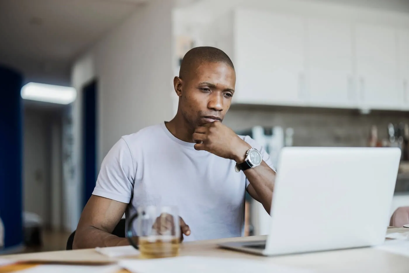 Focused man seated at a table in a home office or kitchen workspace, looking at a laptop with one hand resting on the chin. Papers and a glass mug sit on the table, with cabinets and countertops visible in the background.
