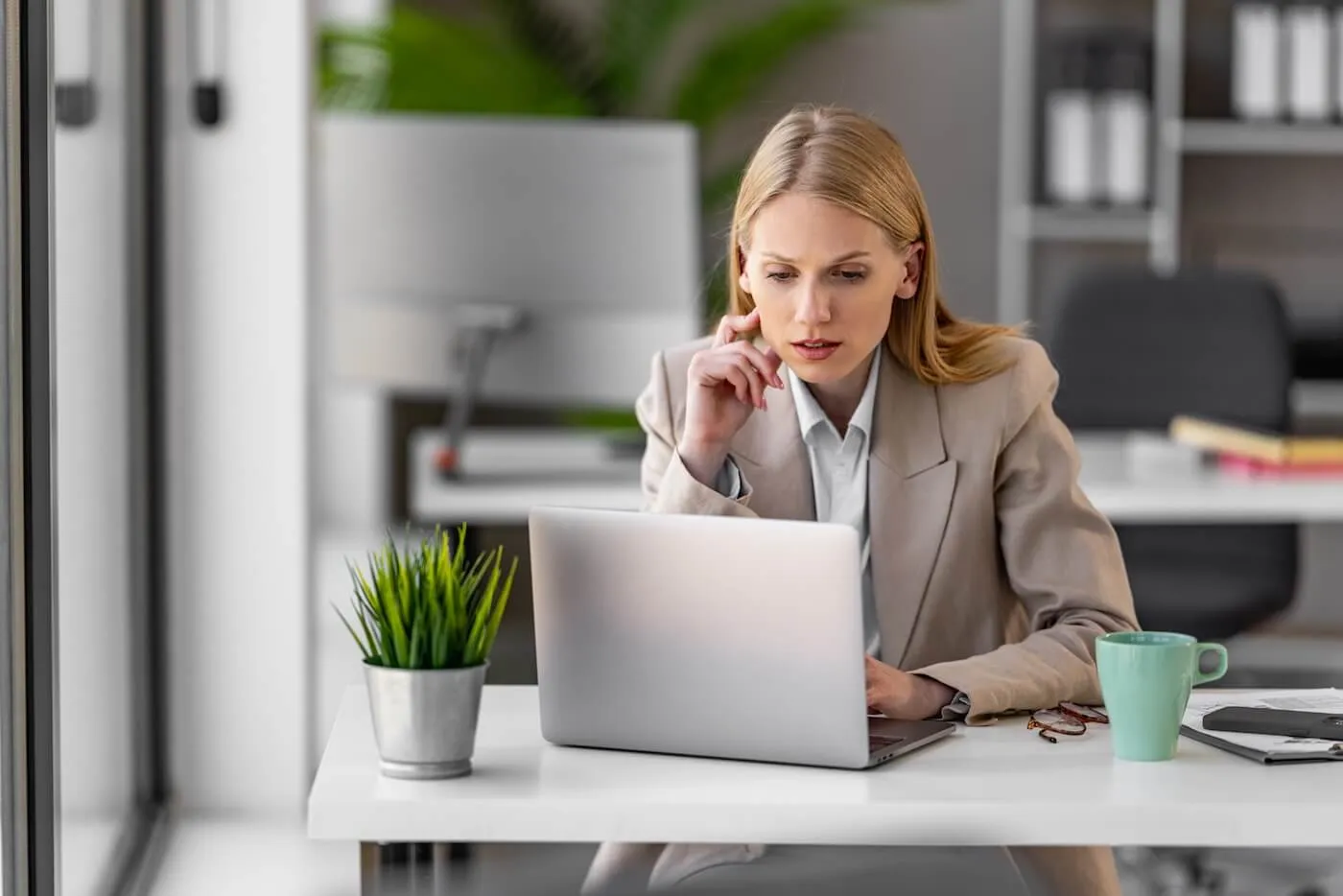 Focused businesswoman seated at a desk using a laptop in a modern office workspace. A coffee mug, notebook, eyeglasses, and potted plant are on the desk, with office furniture and indoor greenery visible in the background.
