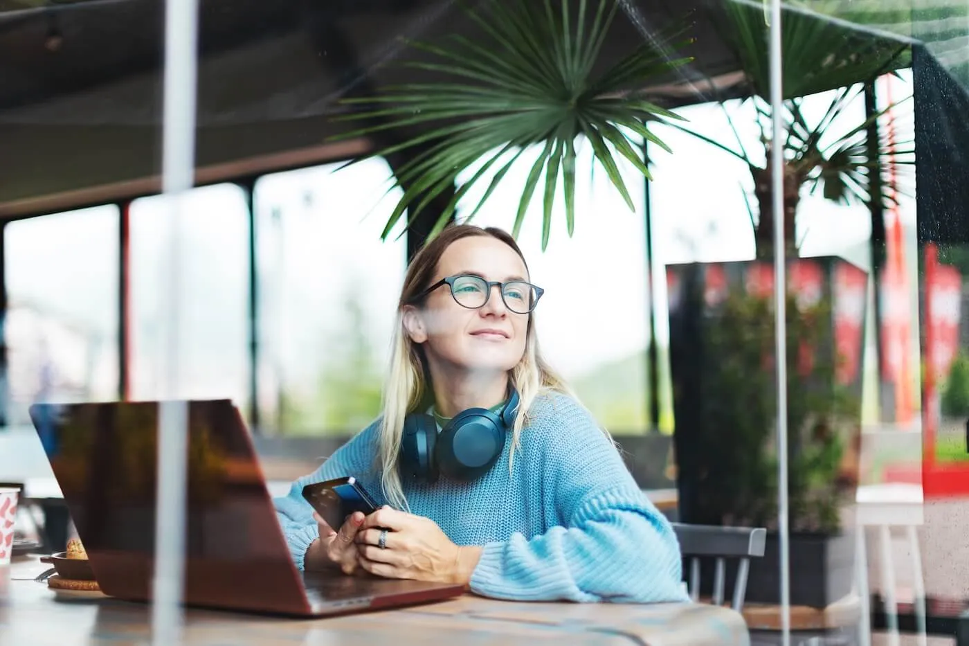 Female freelancer seated at a café table using a laptop and holding a smartphone, with headphones around the neck. The café setting features large windows, potted plants, and natural daylight, illustrating remote work or independent business activity.