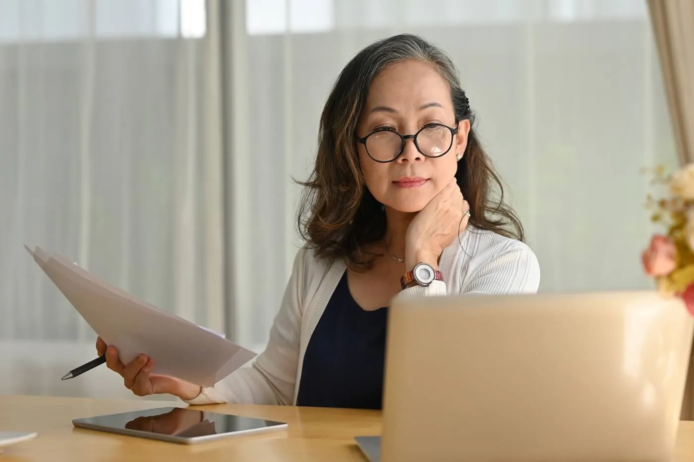 Mature woman sitting at a desk at home, reviewing documents while working on a laptop, with a tablet and pen placed on the table