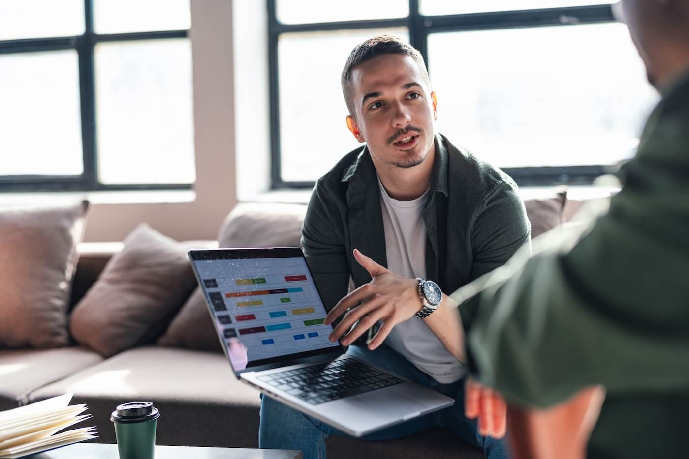 Young man sitting on a couch showing a laptop screen with a colorful chart or dashboard to another person.