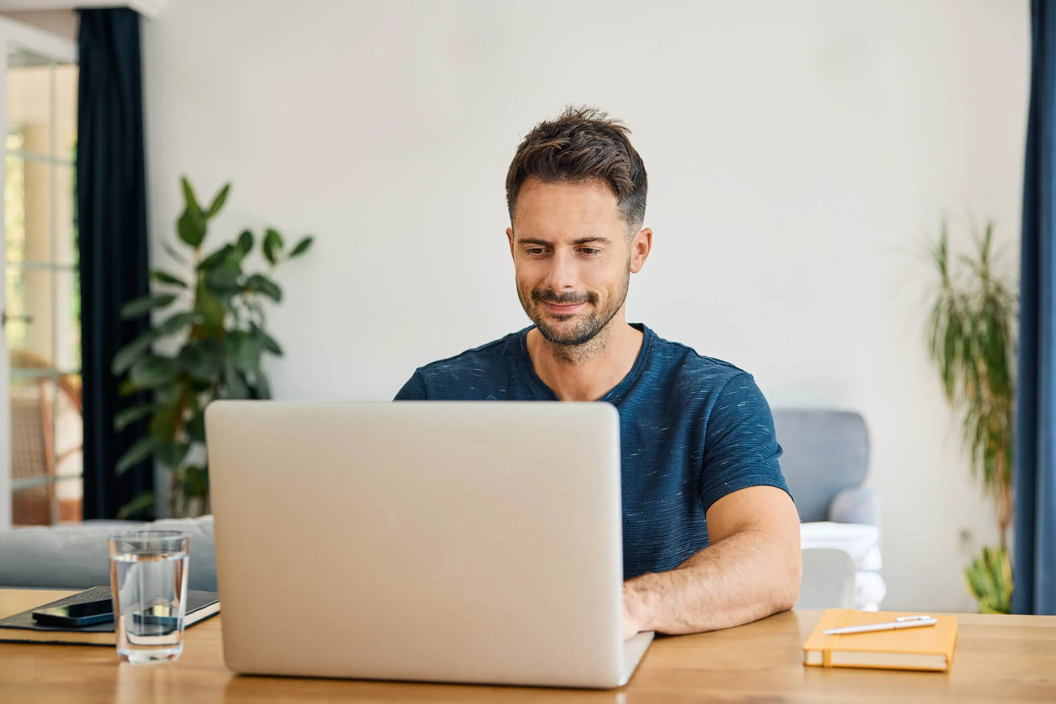 Smiling man sits at a wooden table working on a laptop, with a glass of water, a smartphone, and a notebook placed on the table, in a bright home interior with plants and furniture in the background.