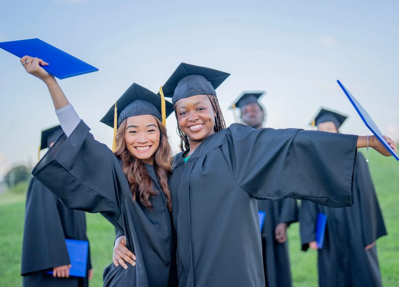 Two female graduates in caps and gowns holding diploma covers during an outdoor commencement ceremony