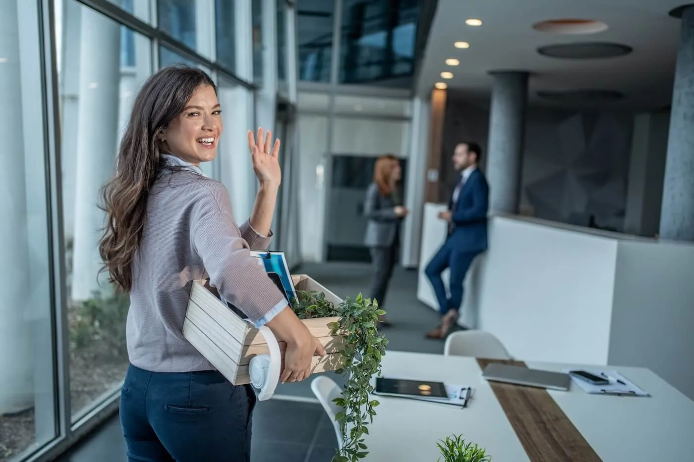 Happy woman carrying a box containing office supplies, folders, headphones, and a plant while waving goodbye and walking through a modern office lobby, with desks, large windows, and coworkers visible in the background.