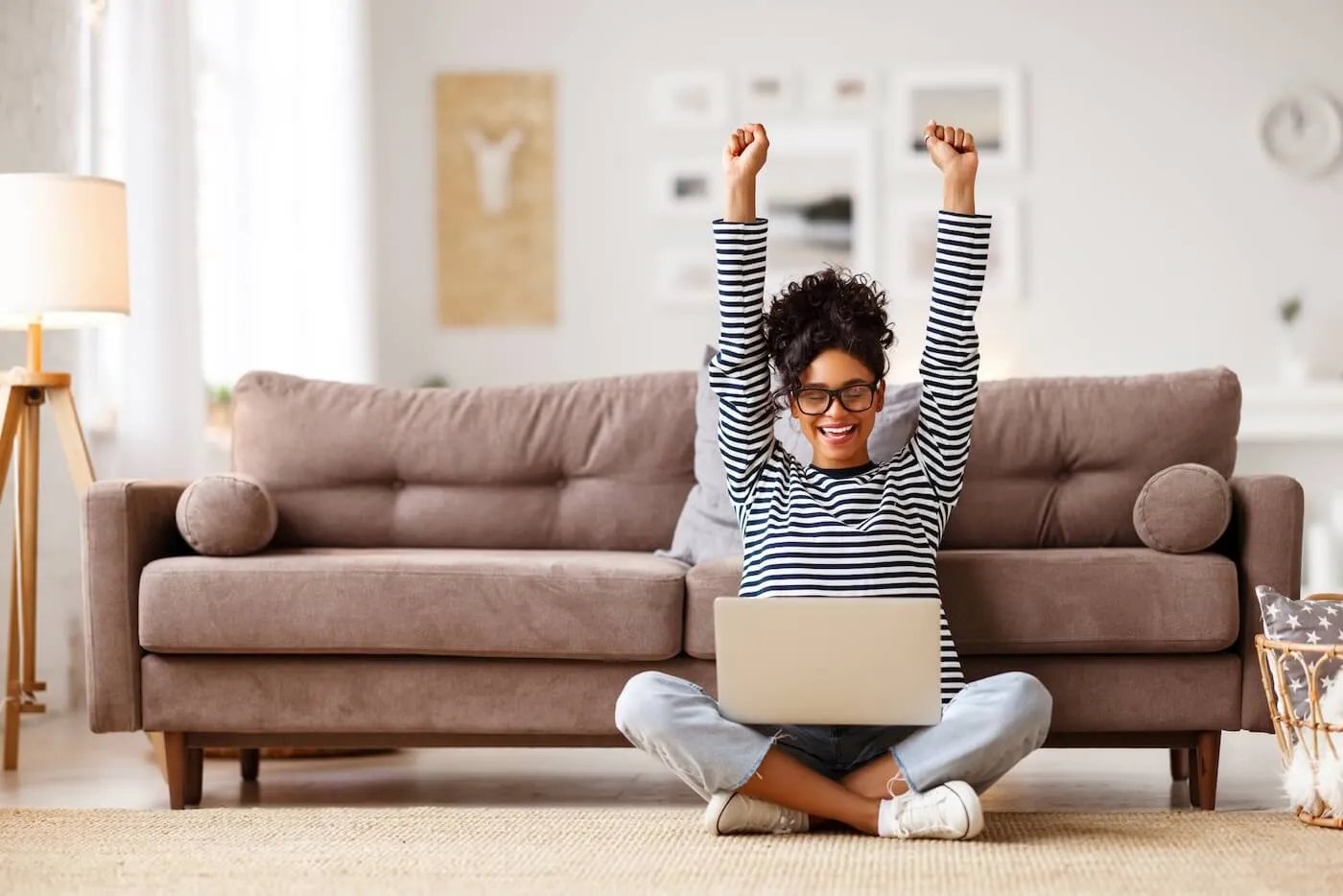 Happy young woman sitting cross-legged on the floor in front of a sofa with a laptop, raising both arms overhead while using the computer in a bright living room. A floor lamp, framed wall art, and home furnishings are visible in the background.