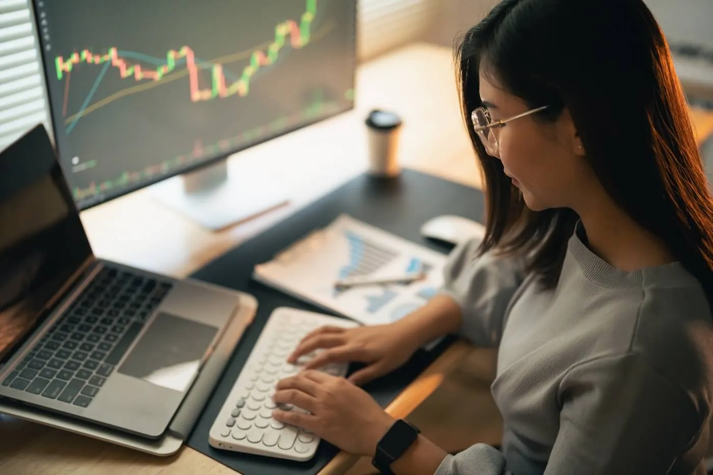 A woman sits at a desk using a keyboard and laptop, with a large monitor displaying financial charts and printed graphs and notes on the table nearby.