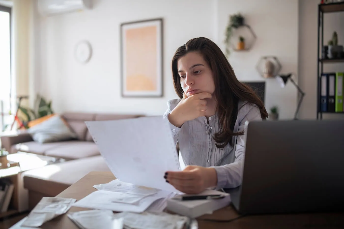 Pensive young woman reviewing a printed document while sitting at the desk with an open laptop and other printouts