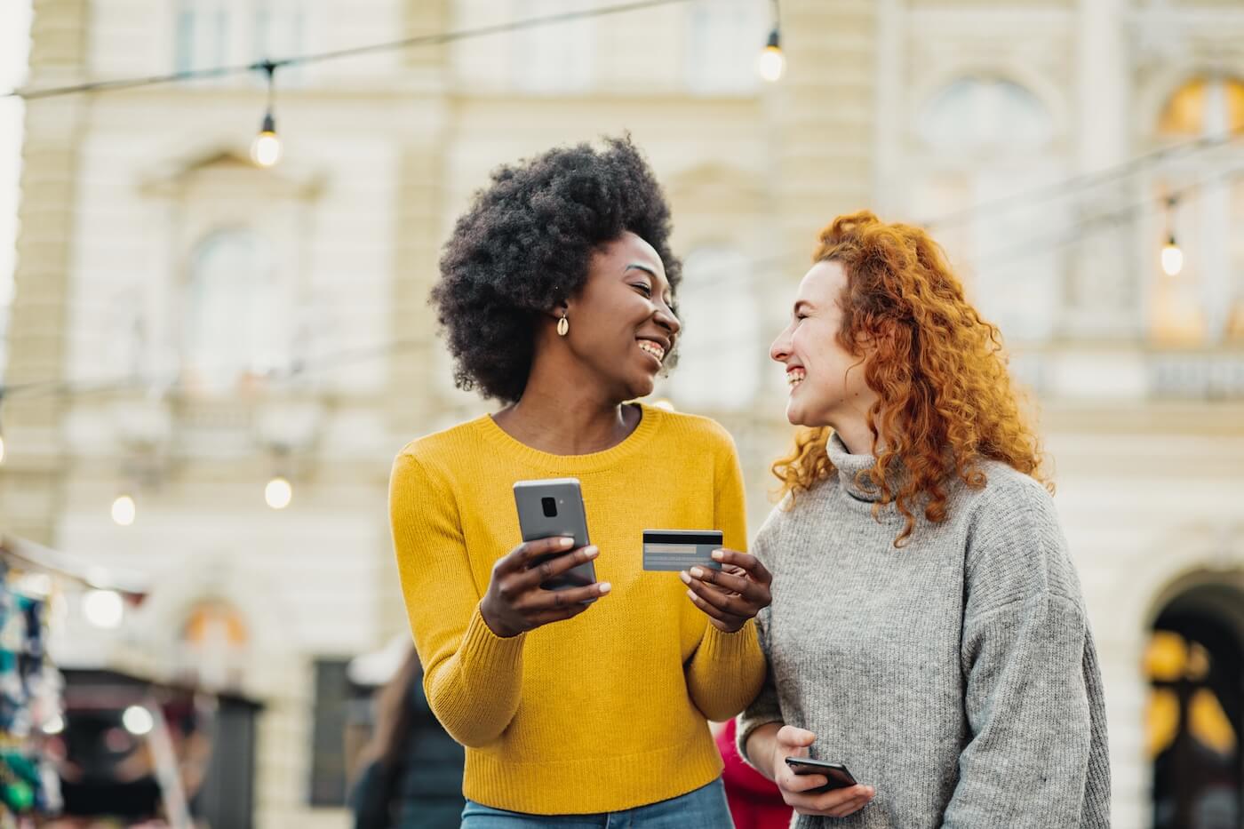 Two happy female friends looking at each other while holding a smartphone and a credit card in a city setting
