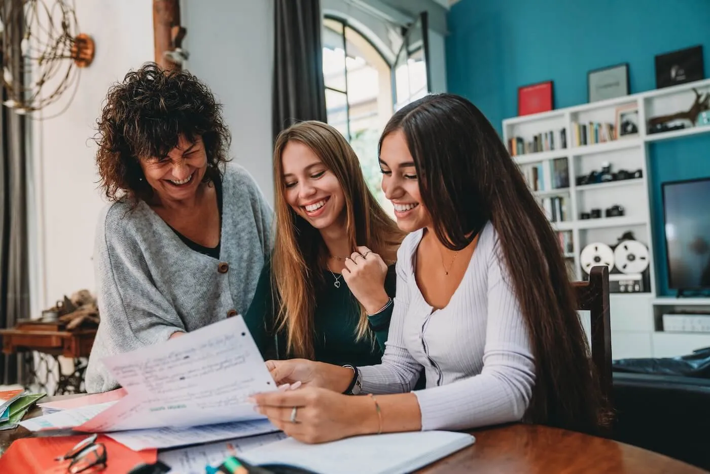Three women sit at a table reviewing a printed document together, with papers, notebooks, and office items spread out in a home setting.