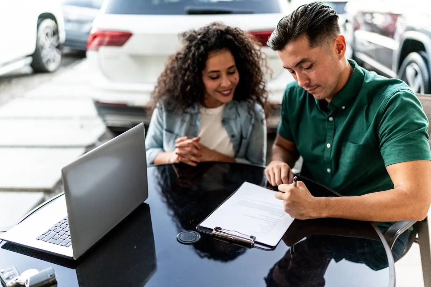 A couple sitting at a glass-topped desk at the car dealership, signing a document, with an open laptop beside them
