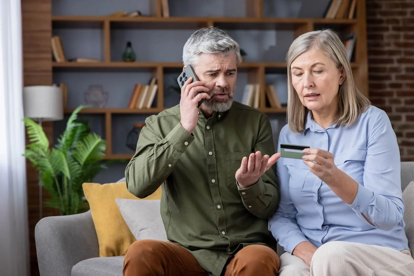 Concerned mature couple sitting on a couch in a living room, the woman holding a credit card while the man talks on a smartphone, with bookshelves, plants, and home décor visible in the background.