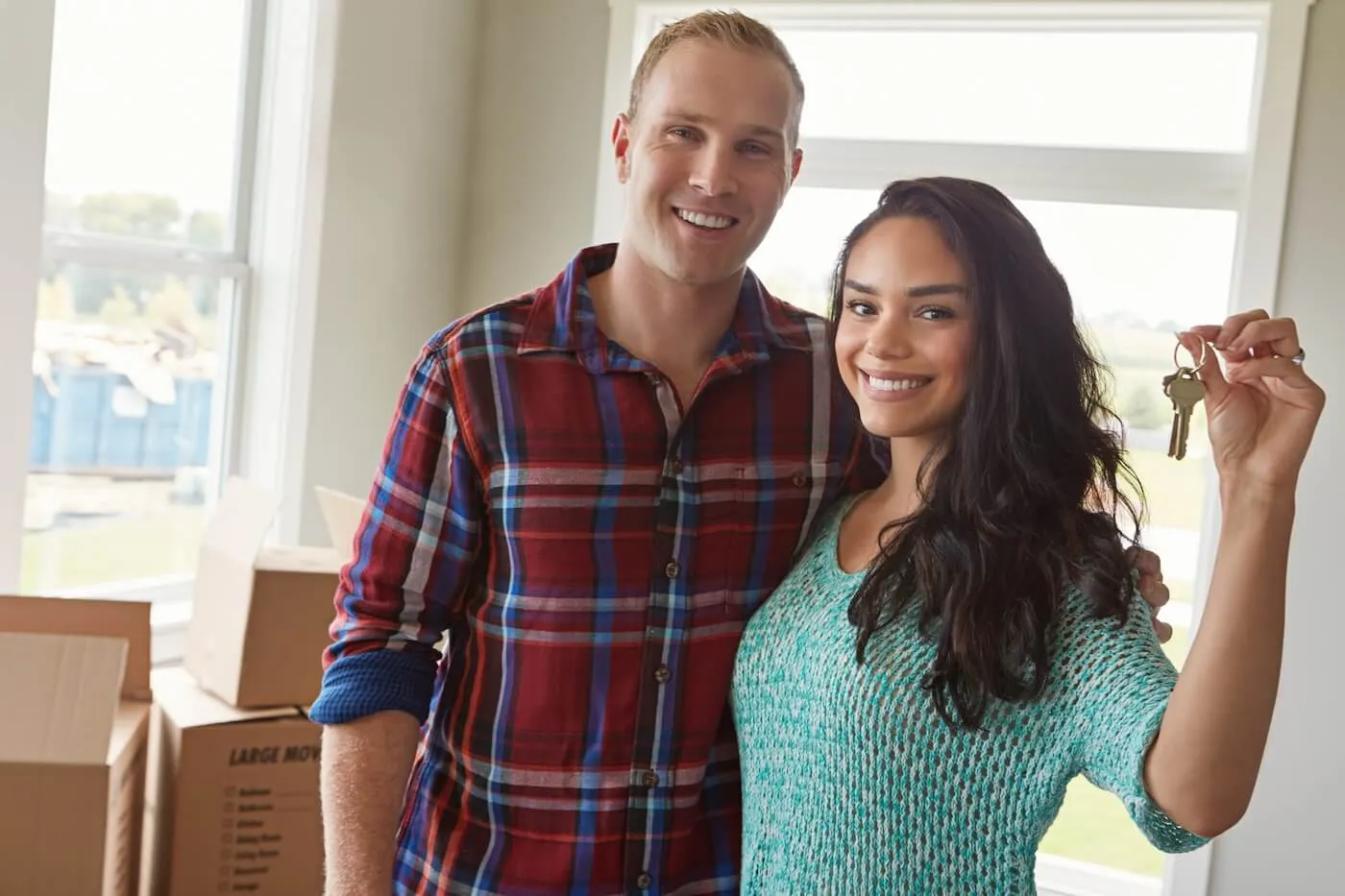 Happy young couple stands inside a home holding a set of keys, with moving boxes visible nearby.