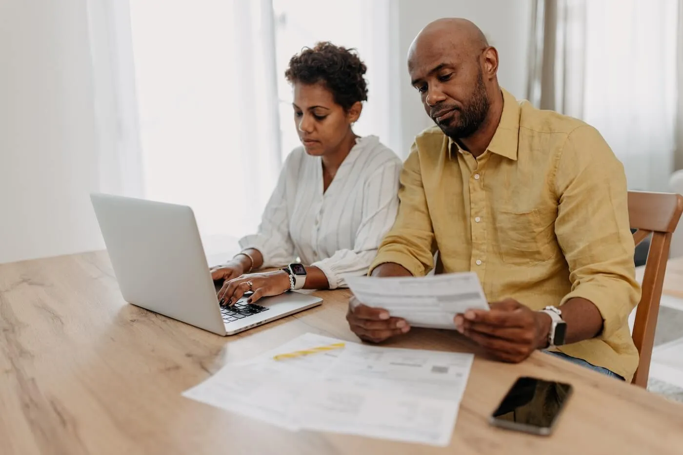 Focused couple sitting at a table, reviewing paperwork, with the woman using a laptop and the man holding a document.