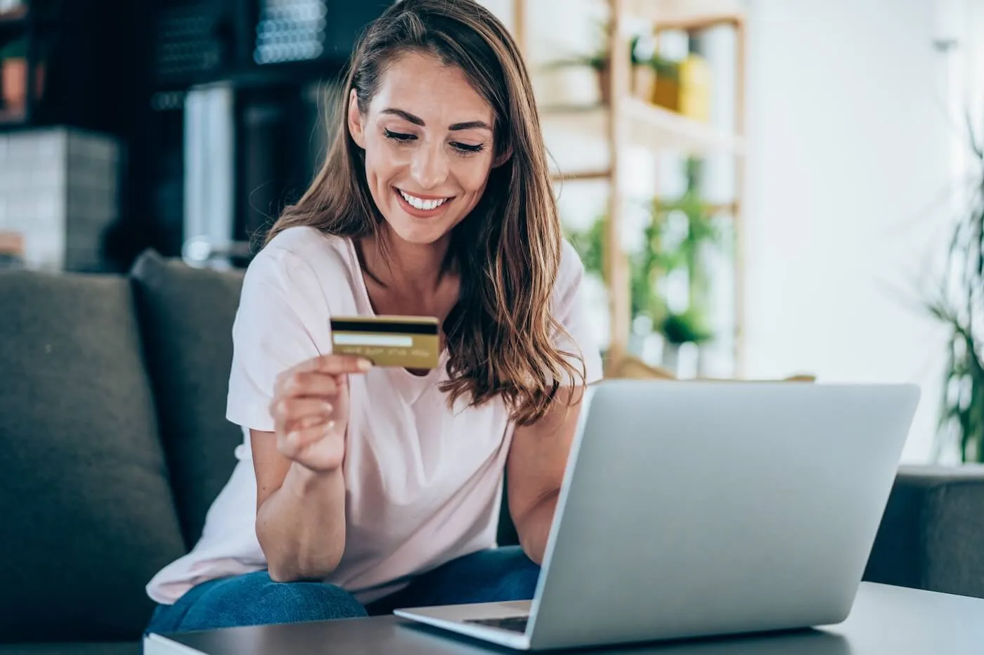 Smiling woman sitting on a couch holding a credit card while using a laptop in a bright living room