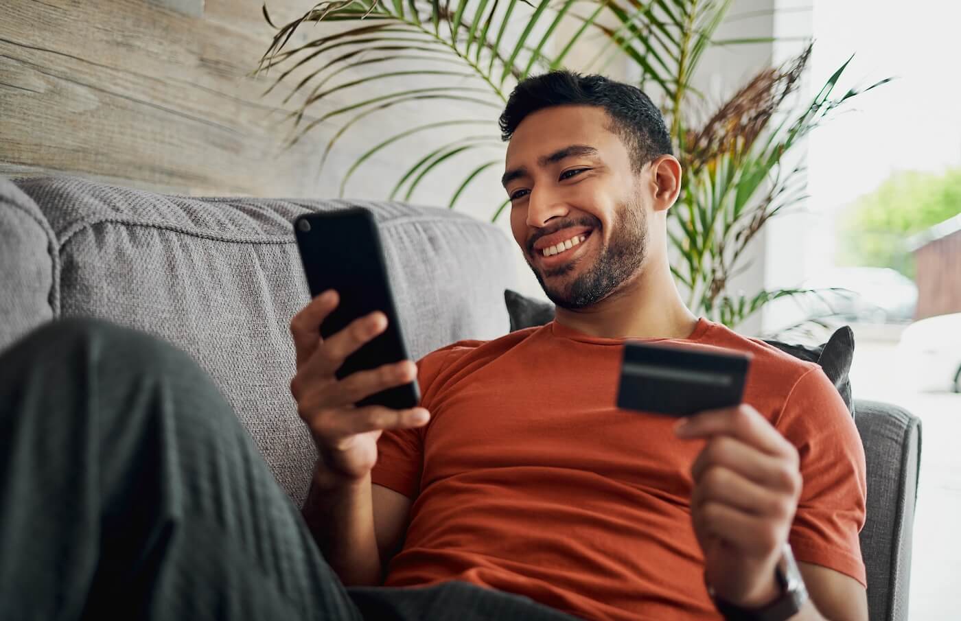 Smiling man lying on a couch holding a smartphone in one hand and a credit card in the other, with indoor plants and a living room setting in the background.