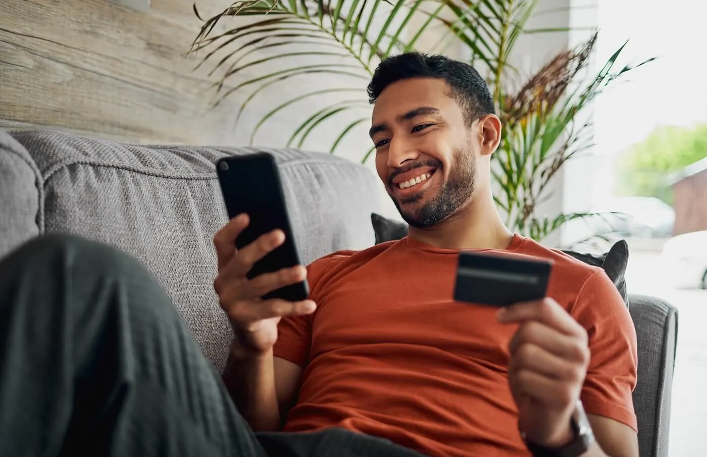 Smiling man lying on a couch holding a smartphone in one hand and a credit card in the other, with indoor plants and a living room setting in the background.