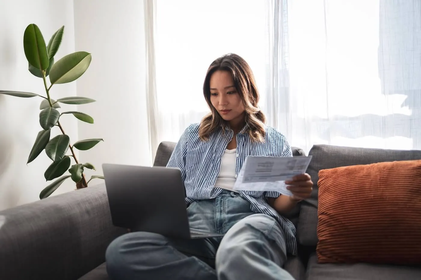 Young woman sitting on a couch using a laptop and holding a document in a bright living room.