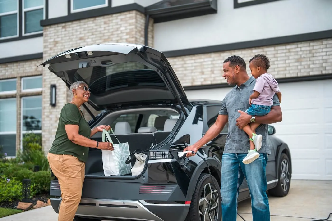 Three people outside a home near an electric vehicle, with the woman loading bags into the trunk and the man holding a small child while unplugging the car from a charger.