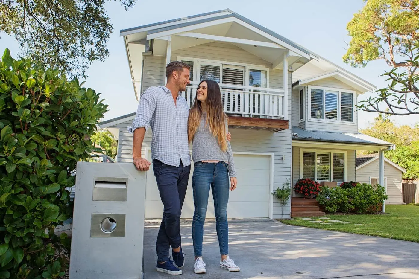 Young couple standing together on a driveway outside a light‑colored house with a balcony and garage, with greenery, shrubs, and a mailbox visible in the foreground.