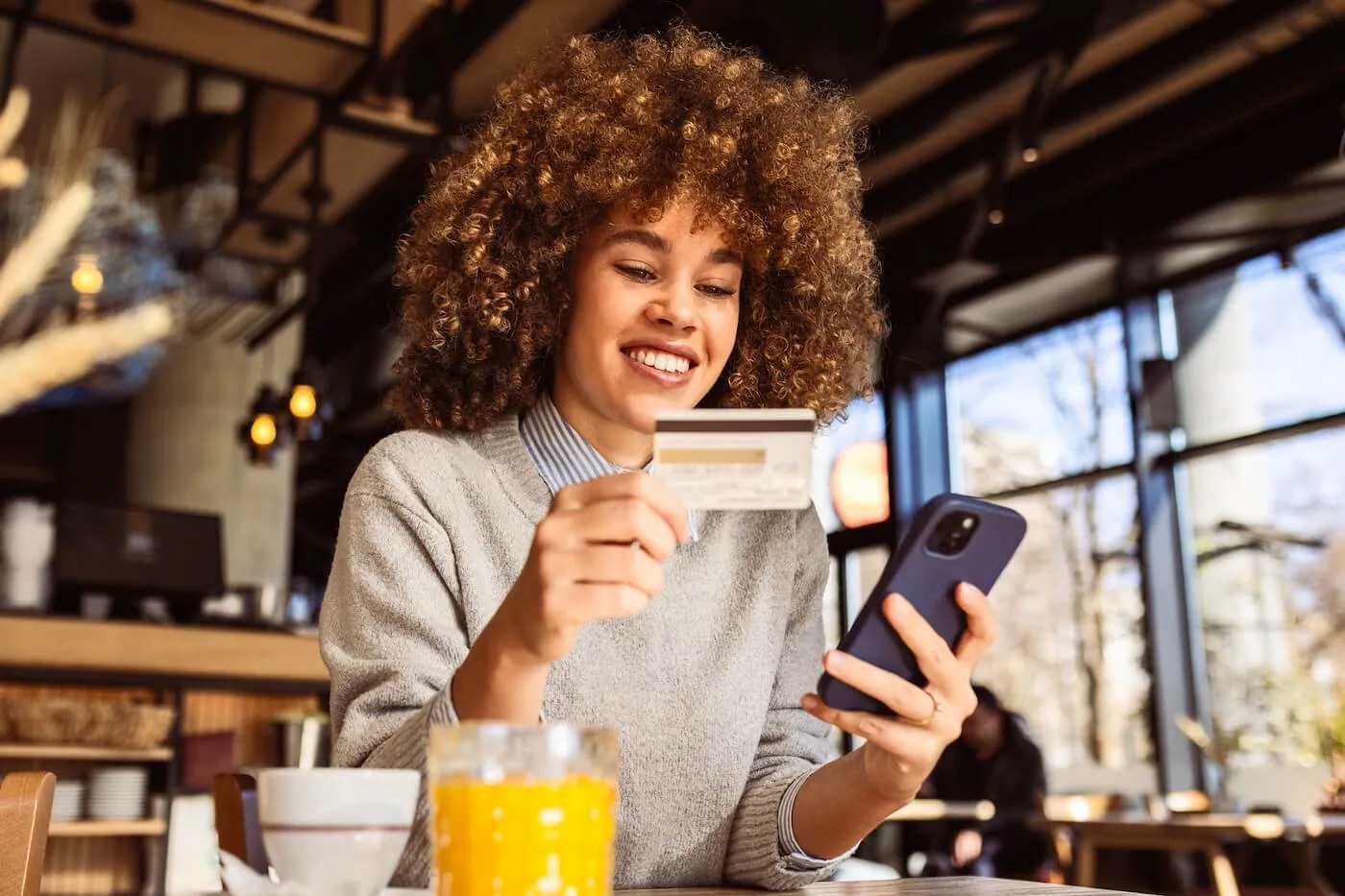 Smiling young woman sits indoors at a café, holding a credit card while looking at a smartphone, with a glass of juice and a cup on the table.