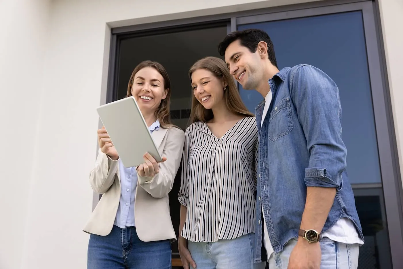 A couple and a female real estate agent standing together outside a modern building entrance, looking at a tablet held by one person, with glass doors and a light-colored exterior wall visible in the background.