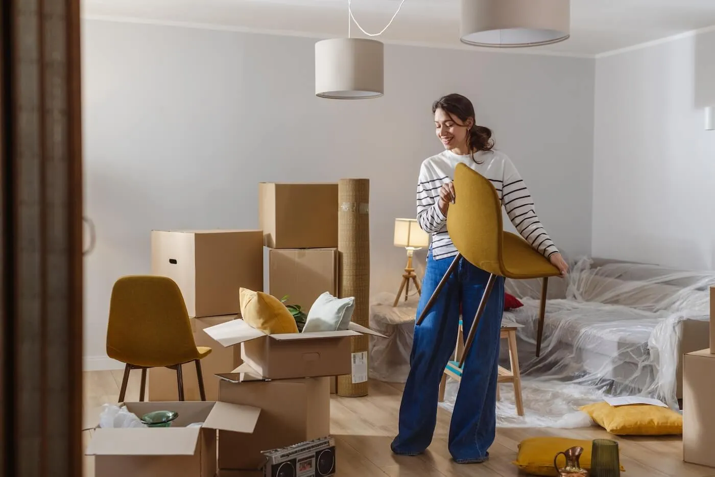 Happy woman stands in a living room holding a chair, surrounded by cardboard boxes, rolled rugs, pillows, and furniture covered in protective wrap.