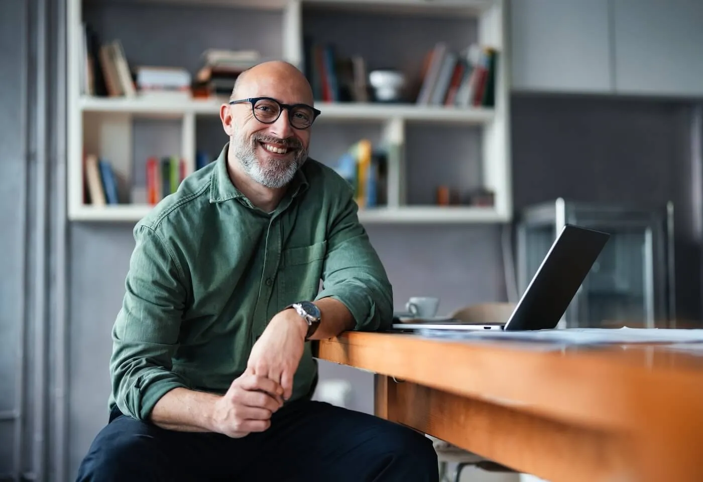Smiling mature man posing for a photo while sitting at a wooden desk in a home office, resting one arm on the tabletop with an open laptop, with bookshelves, books, and office items visible in the background.