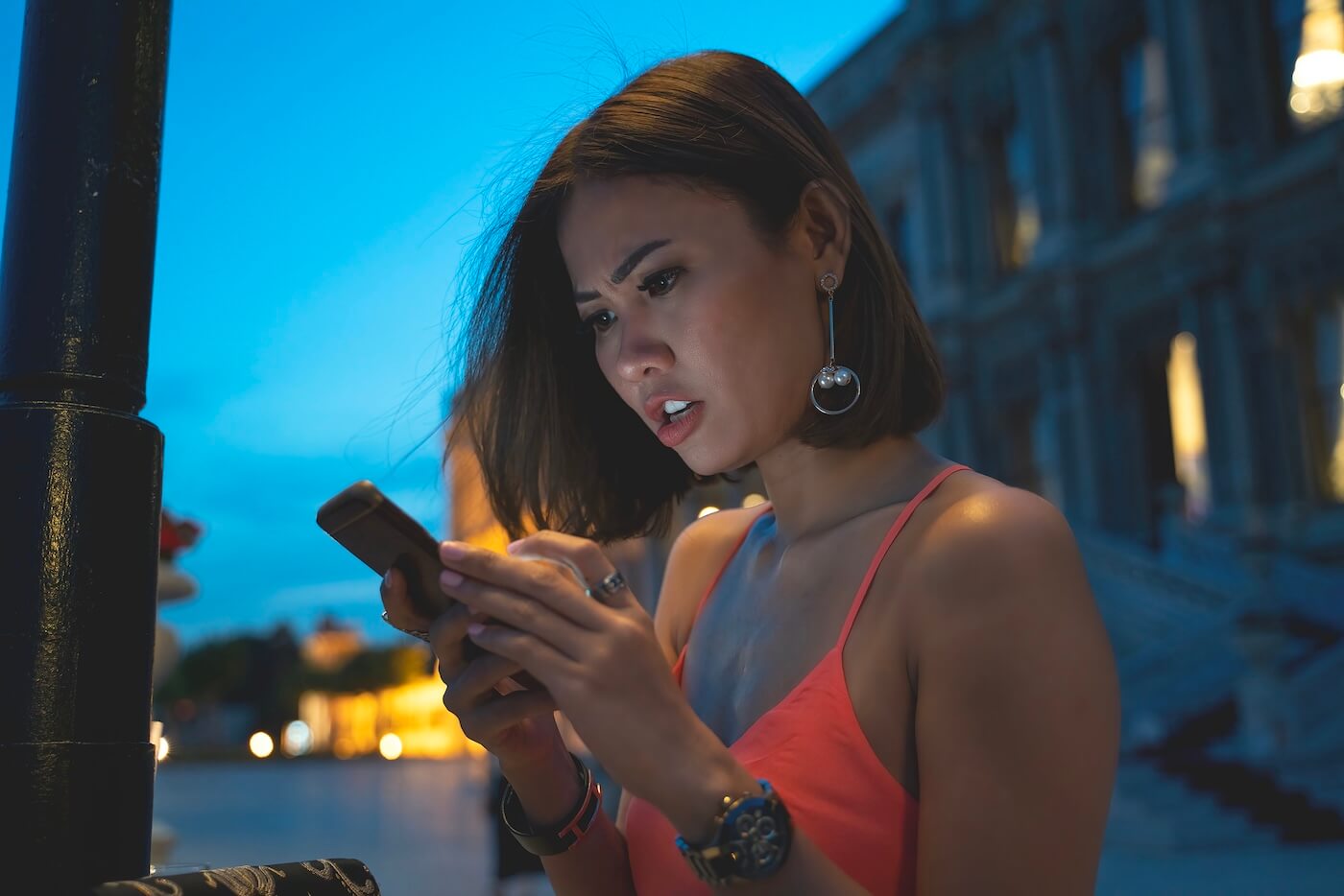 A concerned young woman standing on a dimly lit street at night, looking at her smartphone