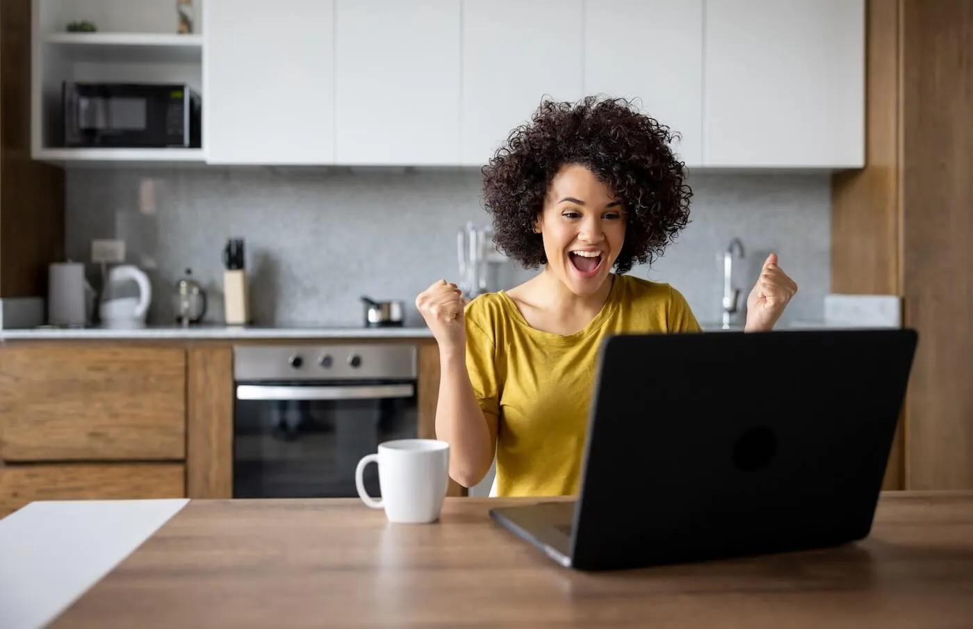 Excited woman looking at her laptop while sitting at the dining table with a mug nearby
