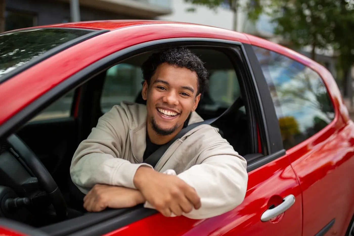 Smiling young man resting their arms on the open window of a red car while sitting in the driver’s seat
