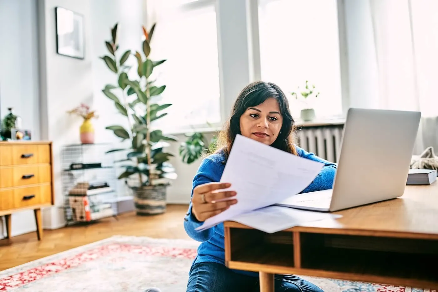 Focused woman sitting on the floor beside a coffee table, reviewing printed documents and using a laptop