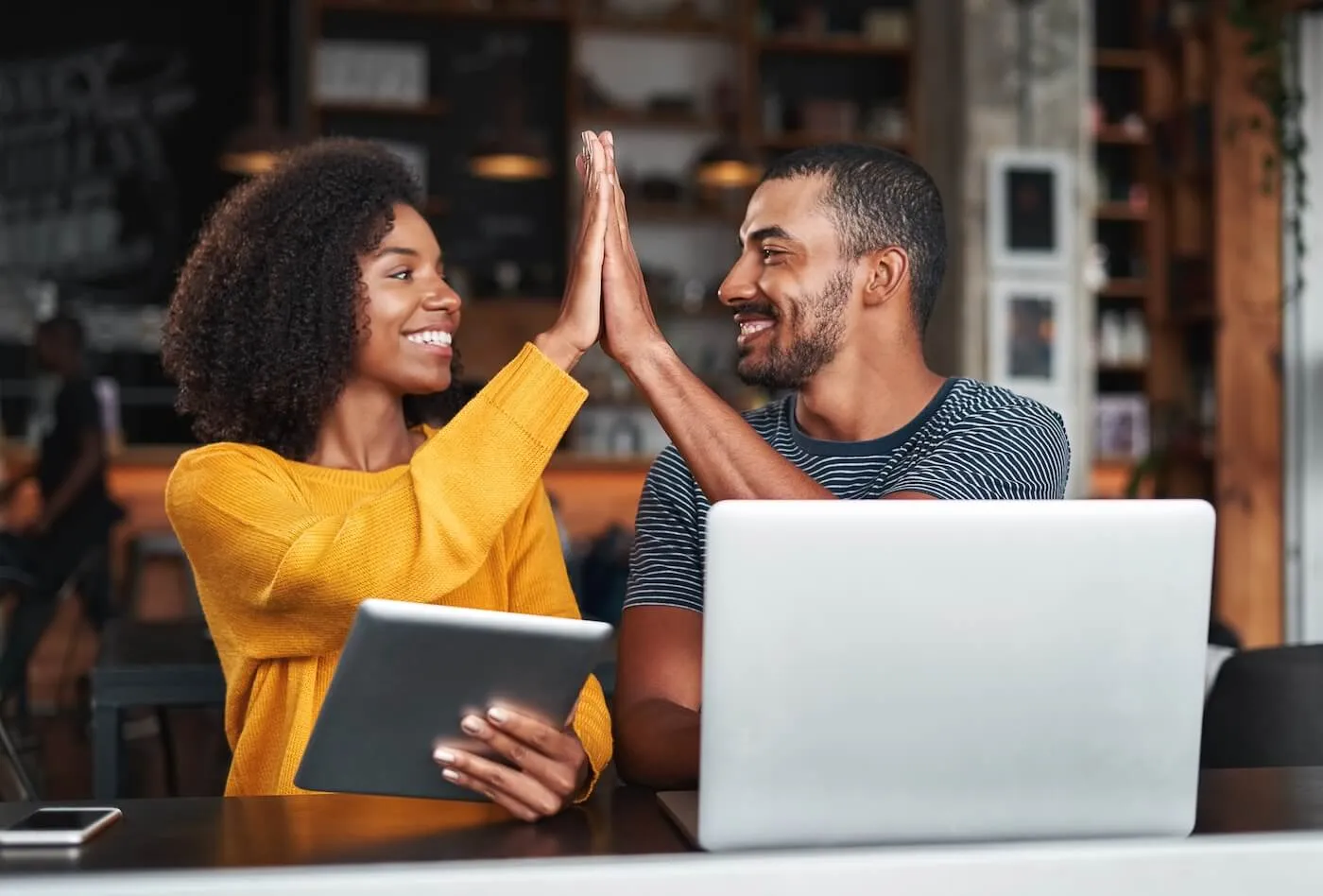 Happy couple sitting at a table in a café, high‑fiving while working with a laptop and a tablet