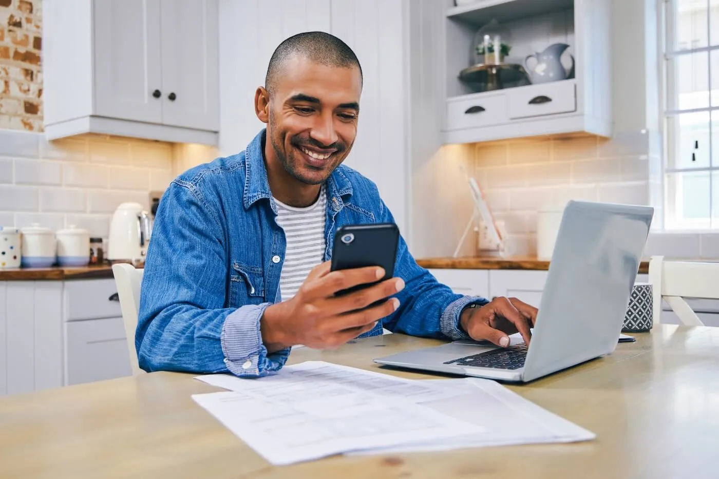Smiling man sitting at a kitchen table using a laptop and smartphone, with documents spread out on the table